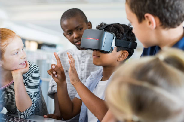 Boy using virtual goggles Mixed race child with vr virtual reality goggles in classroom with his friends. Multiethnic pupil having fun with virtual reality headset at elementary school. Happy boy gesturing while using VR headset in classroom. simulator stock pictures, royalty-free photos & images