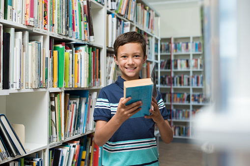 Happy smiling boy reading a book in the library at school. Portrait of cute child in a bookstore looking at camera. Confident pupil borrow a textbook from the library.