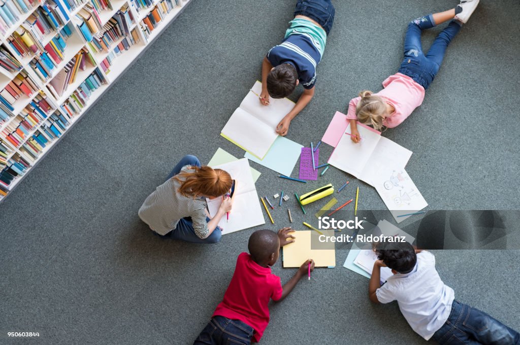 Children drawing at library Elementary children lying on the floor and drawing at library. Top view of five multiethnic boys and girls in daycare house drawing on copybook. High angle view of group of kids drawing with colorful pencils on floor. Child Stock Photo