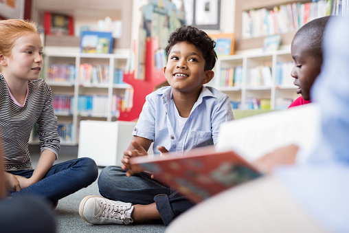 Multiethnic group of kids sitting on floor in circle around the teacher and listening a story. Discussion group of multiethnic children in library talking to woman. Portrait of smiling hispanic boy in elementary school.