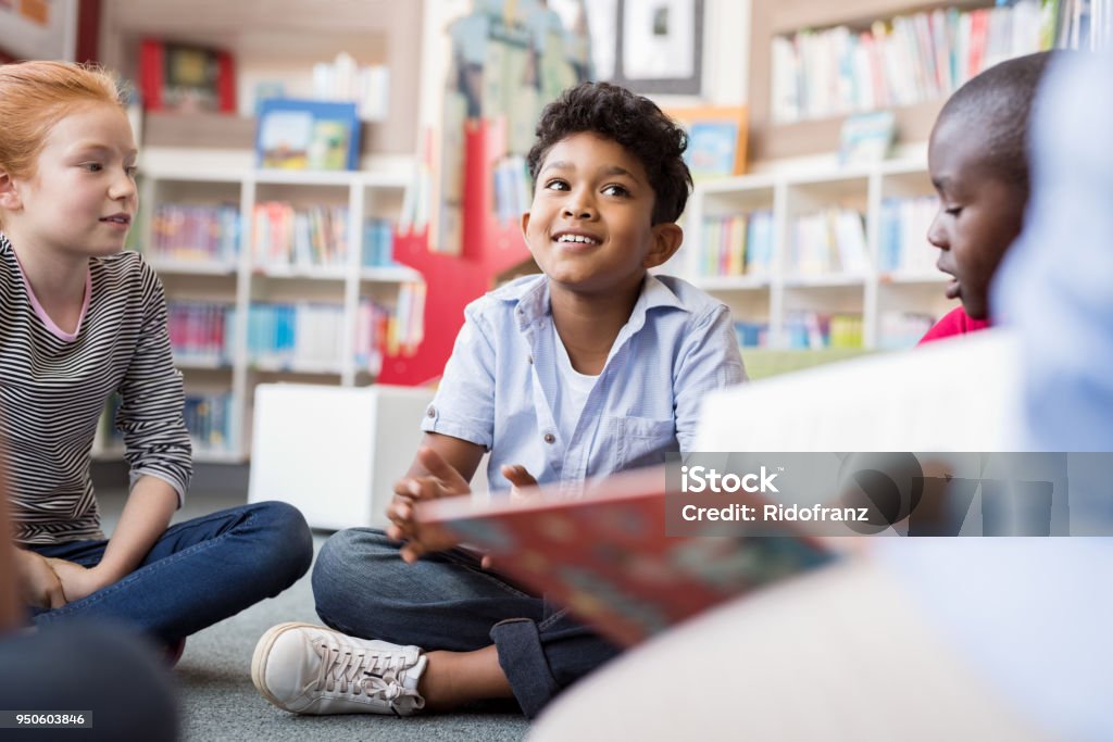 Children listening stories Multiethnic group of kids sitting on floor in circle around the teacher and listening a story. Discussion group of multiethnic children in library talking to woman. Portrait of smiling hispanic boy in elementary school. Child Stock Photo