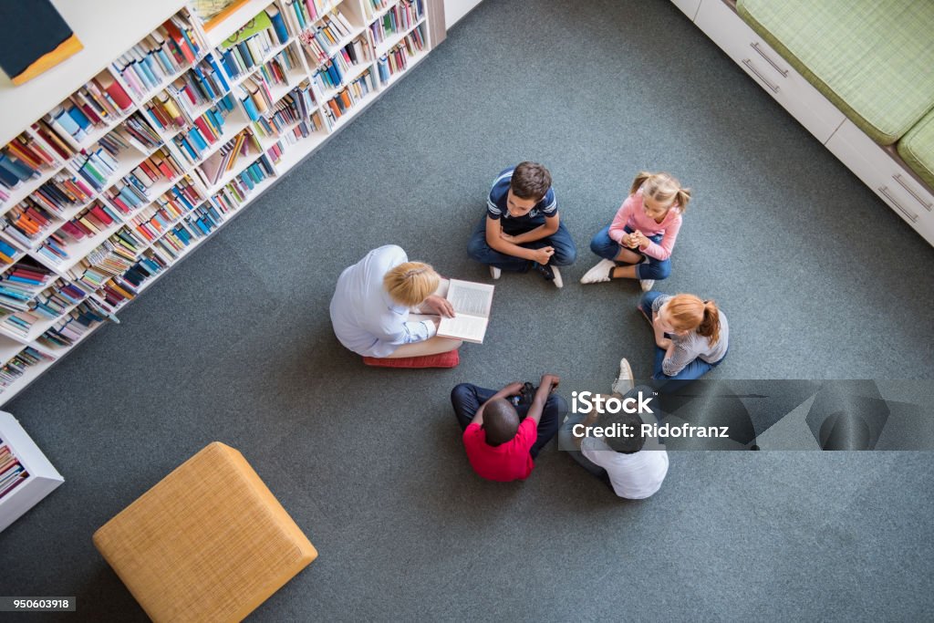 Children listening a fairy tale Teacher reading fairy tales to children sitting in a circle at library. Top view of librarian sitting with five multiethnic children on floor. Teacher reading book to cute girls and young boys at school. Child Stock Photo