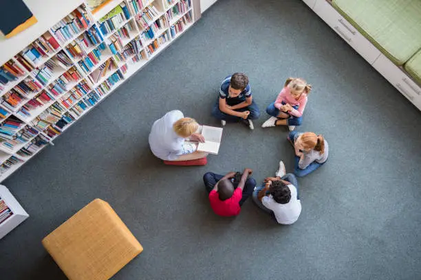 Teacher reading fairy tales to children sitting in a circle at library. Top view of librarian sitting with five multiethnic children on floor. Teacher reading book to cute girls and young boys at school.