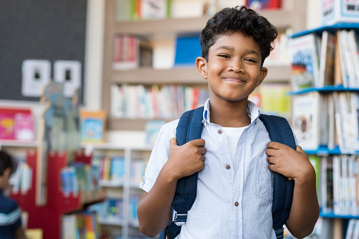 Portrait of smiling hispanic boy looking at camera. Young elementary schoolboy carrying backpack and standing in library at school. Cheerful middle eastern child standing with library background.