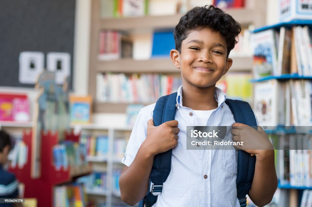 Smiling hispanic boy at school Portrait of smiling hispanic boy looking at camera. Young elementary schoolboy carrying backpack and standing in library at school. Cheerful middle eastern child standing with library background. Child Stock Photo