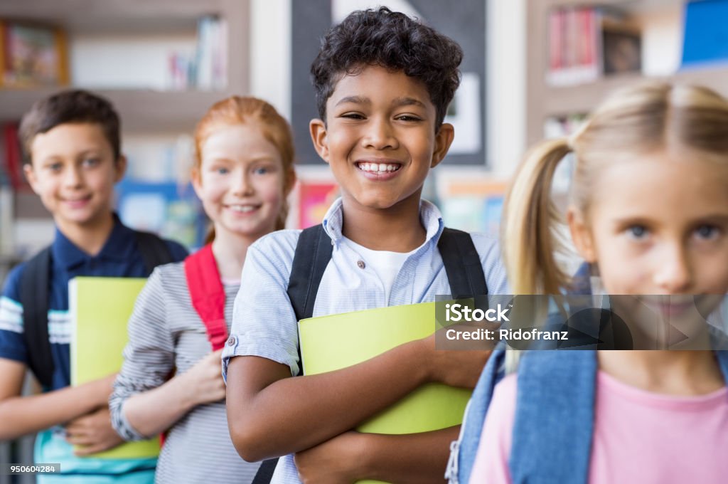 School children standing in a row Portrait of a happy multiethnic children holding books and wearing backpack at primary school. Schoolboys and cute girls in a row holding notebook and looking at camera. Portrait of a elementary child smiling. Child Stock Photo