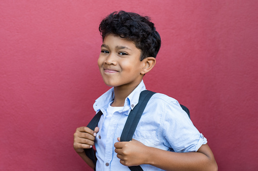 Young hispanic school boy ready with backpack to go to elementary school. Portrait of happy middle eastern schoolboy standing against red background. Indian child looking at camera isolated with copy space.