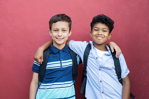 Best children friends standing with hand on shoulder against red background. Happy smiling classmates standing together on red wall after school. Portrait of multiethnic schoolboys enjoying friendship.
