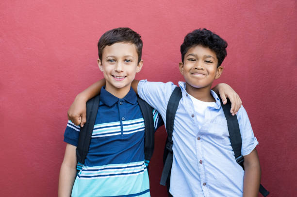 Smiling children friends embracing Best children friends standing with hand on shoulder against red background. Happy smiling classmates standing together on red wall after school. Portrait of multiethnic schoolboys enjoying friendship. preadolescents stock pictures, royalty-free photos & images