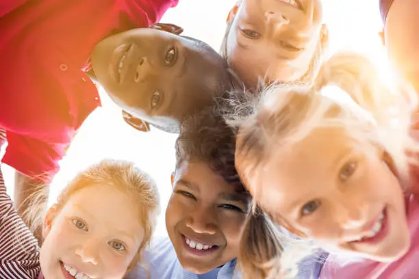 Portrait of happy kids in circle looking down and embracing. Group of five multiethnic friends outdoor looking at camera and smiling. Closeup face of smiling children looking down at the camera together at park.