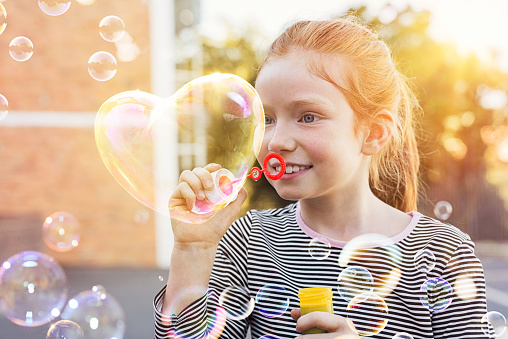 Portrait of funny little girl with red hair blowing heart shaped soap bubble at park. Portrait of cute girl blowing soap bubbles outside the school. Happy child playing with soap bubble at playground.