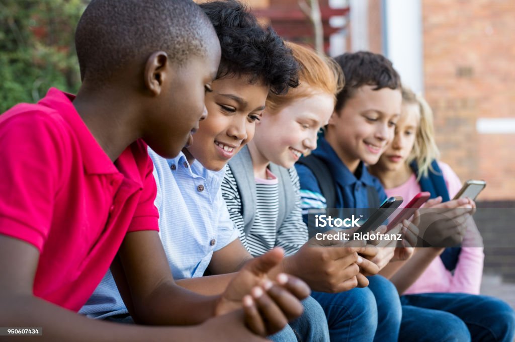 Children using smart phone Pupils using mobile phone at the elementary school during recreation time. Group of multiethnic children sitting in a row and typing a message on smartphone. Young boys and girls playing with cellphone. Child Stock Photo
