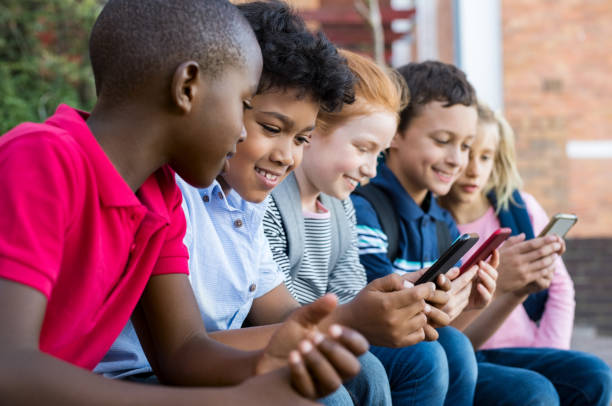 Children using smart phone Pupils using mobile phone at the elementary school during recreation time. Group of multiethnic children sitting in a row and typing a message on smartphone. Young boys and girls playing with cellphone. science and technology kids stock pictures, royalty-free photos & images