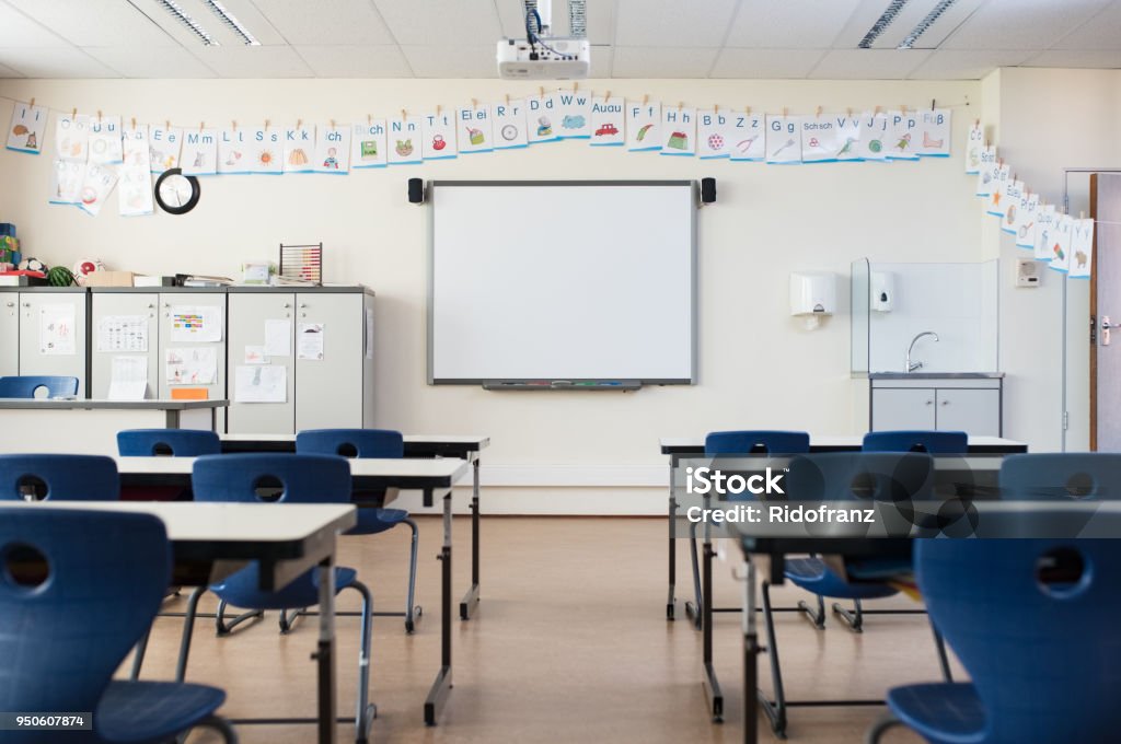 Empty classroom with whiteboard School desk and chairs in empty modern classroom. Empty class room with white board and projector in elementary school. Primary classroom with smartboard and alphabet on wall. Classroom Stock Photo