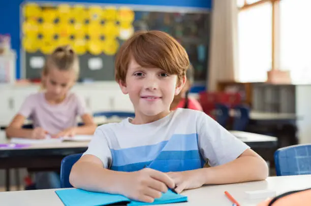 Portrait of happy child with freckles sitting at school desk in class room. Young boy smiling in class and looking at camera with his classmates in background. Pupil with red hair writing on notebook at elementary school.
