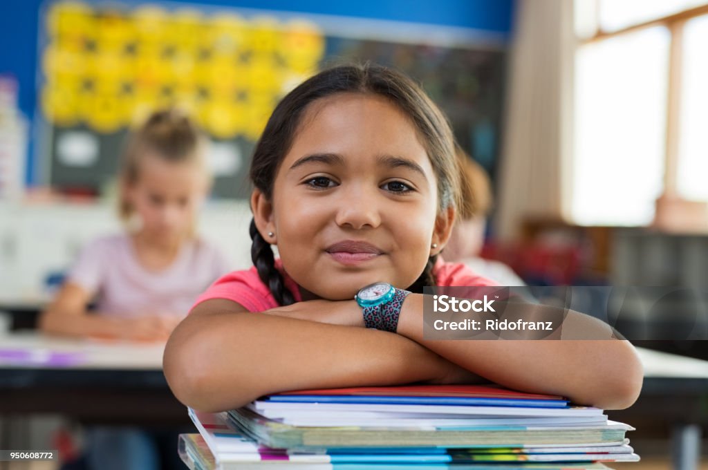 Hispanic girl with chin on books Portrait of cute little schoolgirl leaning on stacked books in classroom. Happy young latin girl in casual keeping chin on notebooks. Closeup face of smiling girl at elementary school. Child Stock Photo