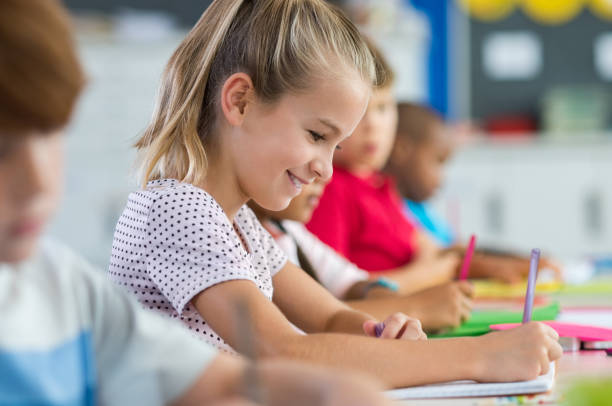 Student girl writing on textbook Smiling scholar girl sitting with other children in classroom and writing on textbook. Happy student doing homework at elementary school. Young schoolgirl feeling confident while writing on notebook. primary school exams stock pictures, royalty-free photos & images