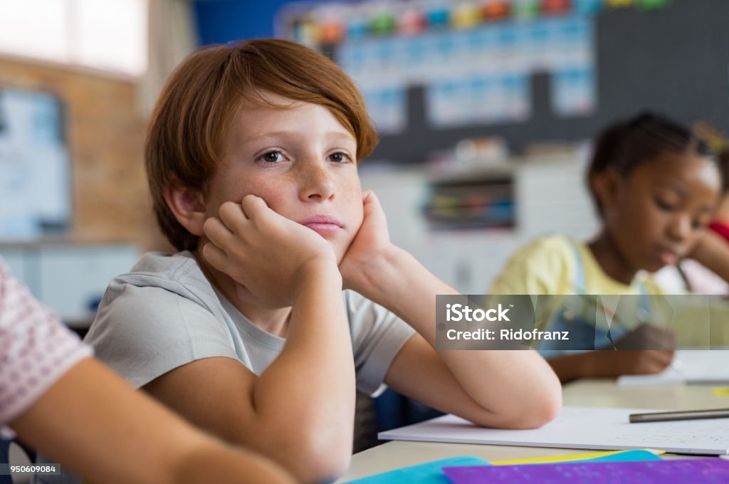Bored school boy in class Tired school boy with hand on face sitting at desk in classroom. Bored schoolchild sitting at desk with classmates in classroom. Frustrated and thoughtful young child sitting and looking up. Child Stock Photo