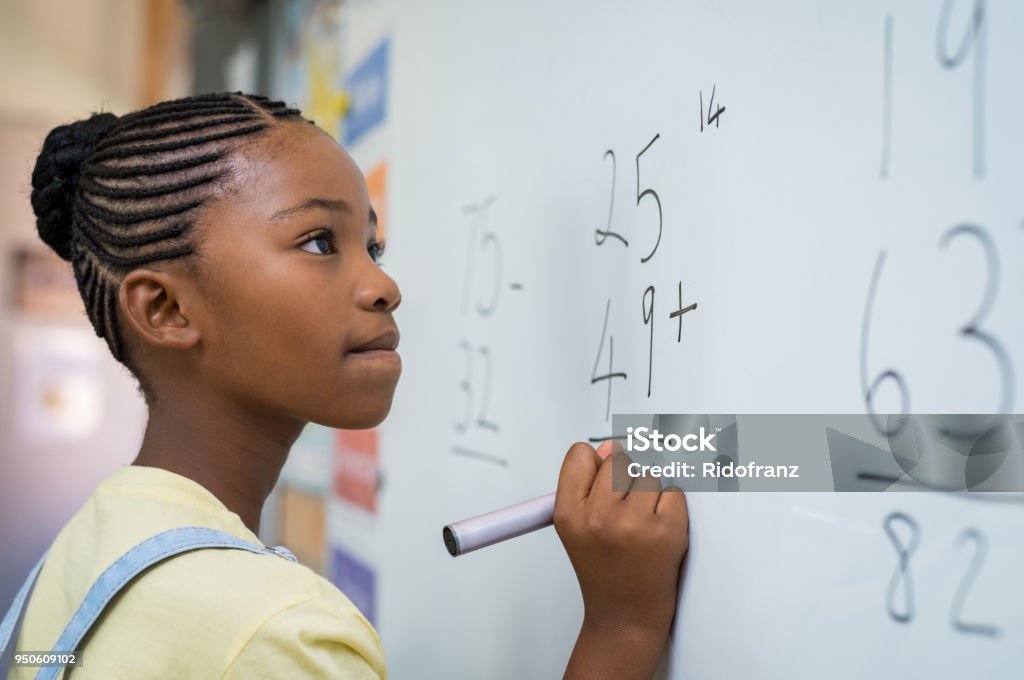 Girl solving mathematical addition Portrait of african girl writing solution of sums on white board at school. Black schoolgirl solving addition sum on white board with marker pen. School child thinking while doing mathematics problem. Mathematics Stock Photo