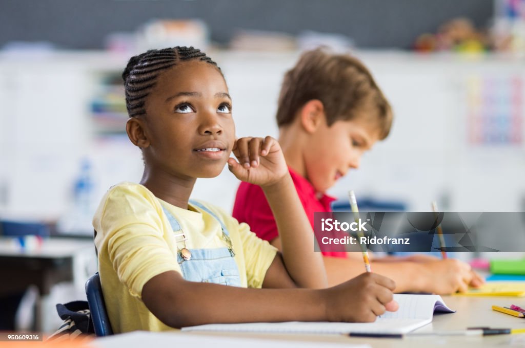 African girl thinking in class Thoughtful elementary school girl sitting at desk and looking up. Thoughtful schoolgirl looking up while sitting at desk during exam with classmate in background. Black scholar thinking about the solution of the exam. Child Stock Photo