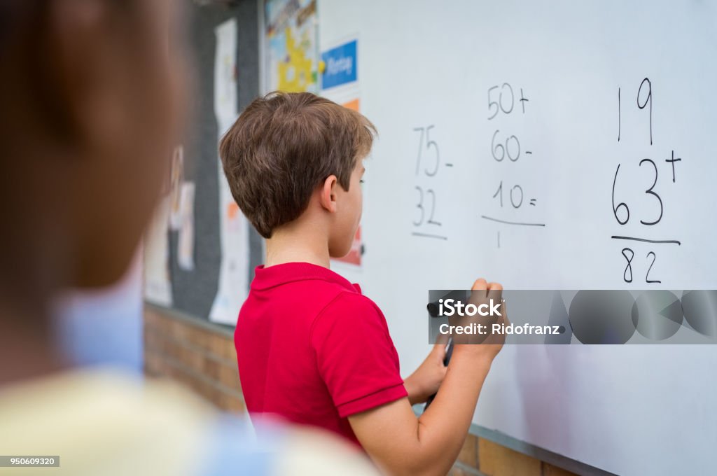 Boy solving math problem Rear view of young boy solving addition and subtraction on white board at school. Schoolboy thinking while solving math's sum. Smart child writing the solution of the mathematical operation in classroom. Mathematics Stock Photo