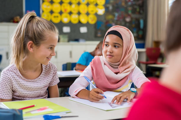 Muslim girl with her classmate Young arab girl with hijab doing exercise with her bestfriend at international school. Asian muslim school girl sitting near her classmate during lesson. Multiethnic elementary students in classroom. hijab stock pictures, royalty-free photos & images