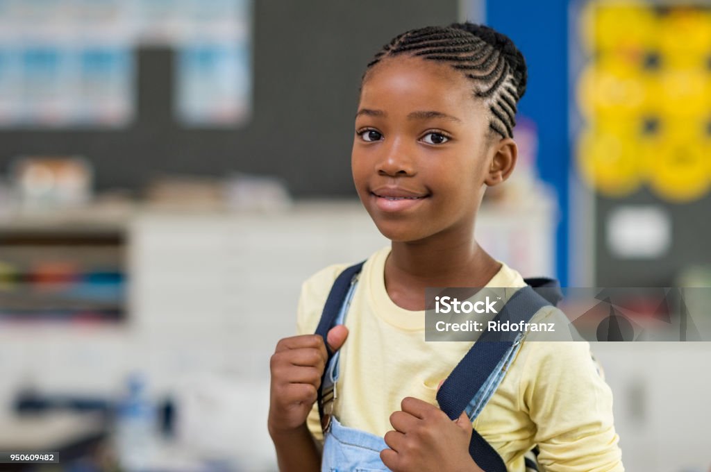 Girl wearing backpack at school African young girl with blue backpack looking at camera. Pretty and satisfied black schoolgirl with rucksack smiling in class. Portrait of beautiful school girl standing in classroom. Child Stock Photo