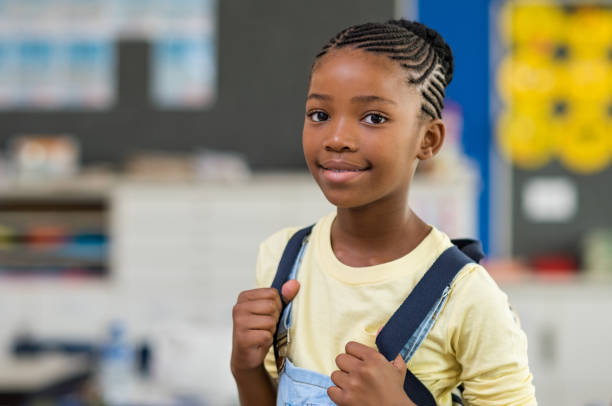 Girl wearing backpack at school African young girl with blue backpack looking at camera. Pretty and satisfied black schoolgirl with rucksack smiling in class. Portrait of beautiful school girl standing in classroom. 8 9 years photos stock pictures, royalty-free photos & images