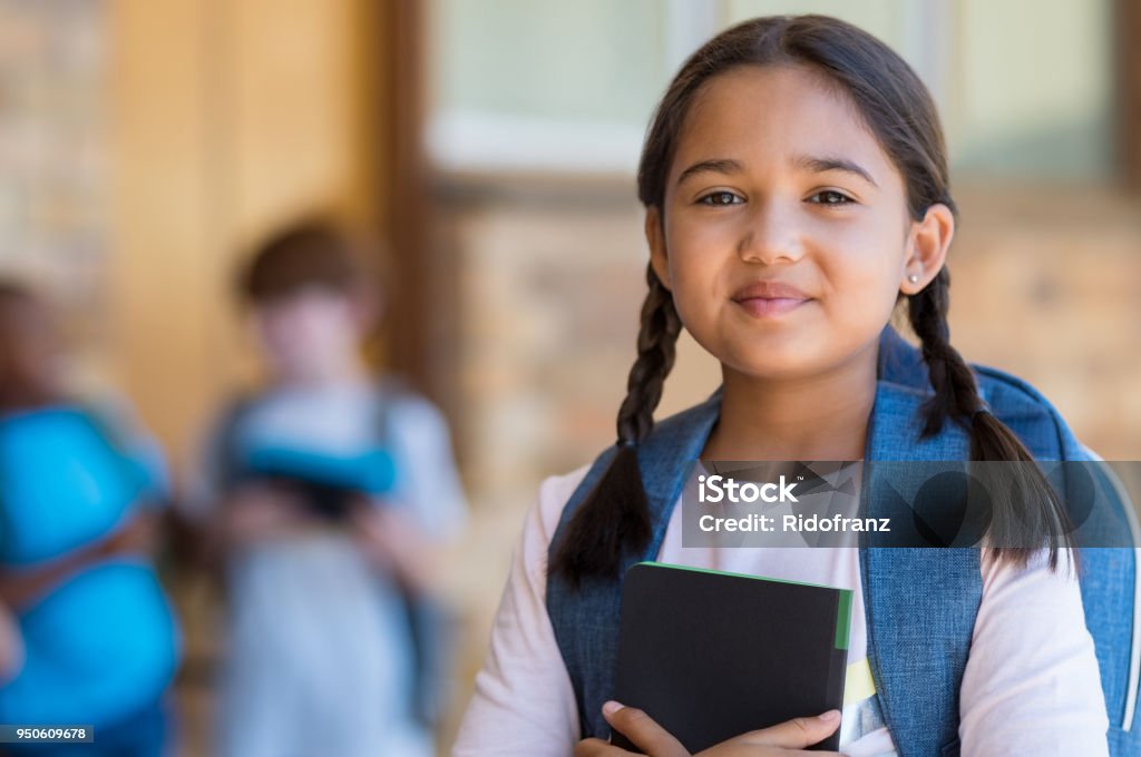 Elementary girl at school Smiling student girl wearing school backpack and holding exercise book. Portrait of happy asian young girl outside the primary school. Closeup face of smiling hispanic schoolgirl looking at camera. Child Stock Photo