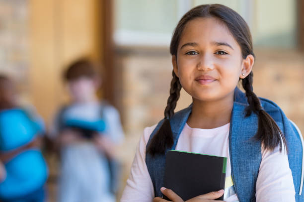 Elementary girl at school Smiling student girl wearing school backpack and holding exercise book. Portrait of happy asian young girl outside the primary school. Closeup face of smiling hispanic schoolgirl looking at camera. smirk stock pictures, royalty-free photos & images