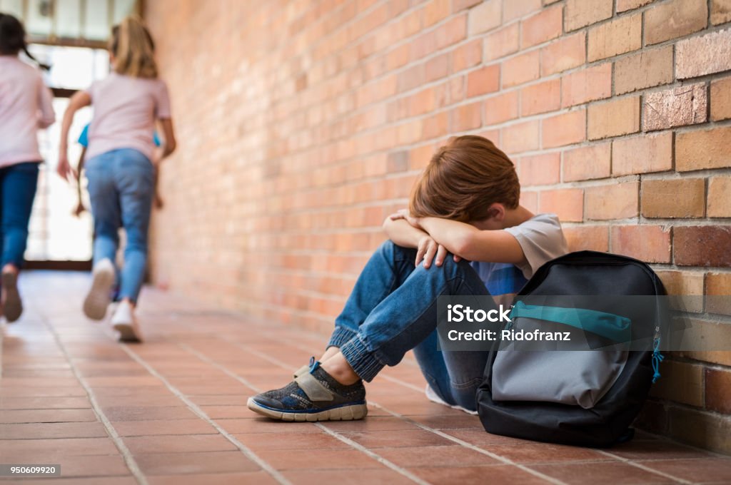 Bullying at school Little boy sitting alone on floor after suffering an act of bullying while children run in the background. Sad young schoolboy sitting on corridor with hands on knees and head between his legs. Bullying Stock Photo