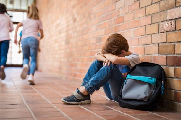 Bullying at school Little boy sitting alone on floor after suffering an act of bullying while children run in the background. Sad young schoolboy sitting on corridor with hands on knees and head between his legs. child sadness stock pictures, royalty-free photos & images