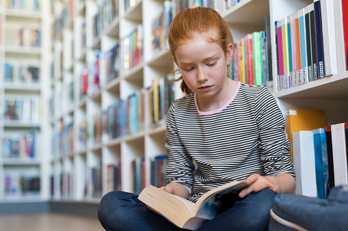 Elementary schoolgirl reading a book at the library. Cute young girl with red hair sitting on floor and reading a book. Diligent pupil concentrated in reading book.