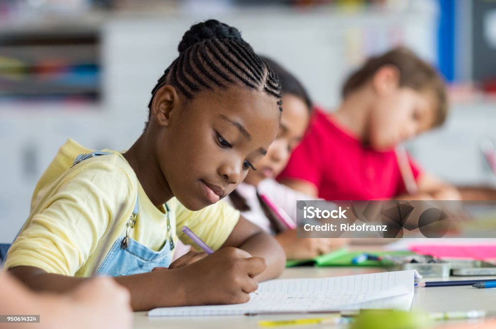 School girl writing in class Cute pupil writing at desk in classroom at the elementary school. Student girl doing test in primary school. Children writing notes in classroom. African schoolgirl writing on notebook during the lesson. Child Stock Photo