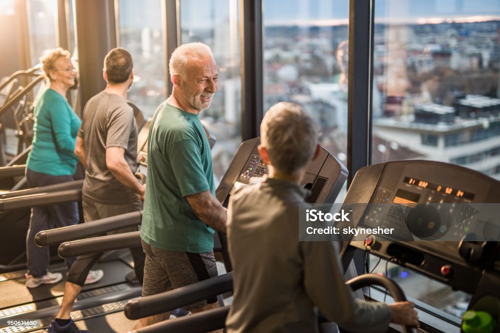 Happy athletic seniors talking while exercising on treadmills in a health club. Four athletic seniors people having a sports training on treadmills in a gym. Focus is on man in green shirt talking to woman next to him. Active Lifestyle Stock Photo