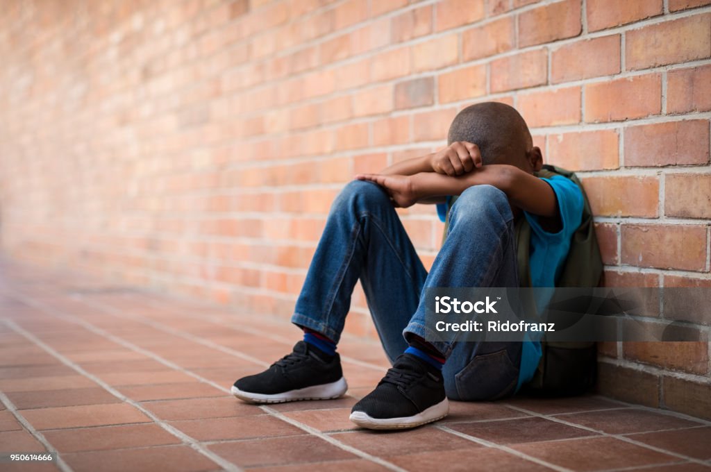 Young sad boy at school Young boy sitting alone with sad feeling at school. Depressed african child abandoned in a corridor and leaning against brick wall. Bullying, discrimination and racism concept at school with copy space. Child Stock Photo