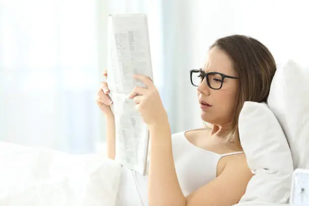 Girl wearing eyeglasses having problems to read a newspaper lying on a bed at home