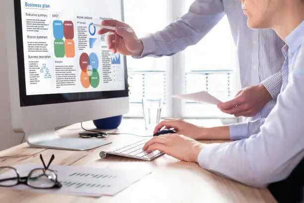 Mixed team of consultants working on a Business Plan report on computer screen with executive summary, strategy information data, SWOT, market analysis, and financial statement, consulting office