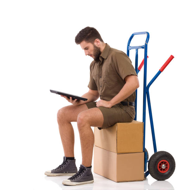Male Messenger Is Sitting On Stack Of Boxes And Looking At Digital Tablet Male Messenger Is Sitting On A Stack Of Boxes, Looking At Digital Tablet And Reading. Full Length Studio Shot Isolated On White. mover delivery person box hand truck stock pictures, royalty-free photos & images