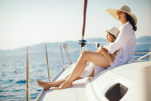 Mother and daughter sitting on yacht deck while sailing over nice calm sea. They both wearing white summer dress and they have hats.