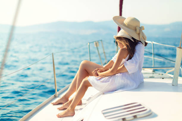 Mother and daughter sitting on yacht deck while sailing over nice calm sea. They both wearing white summer dress and they have hats.
