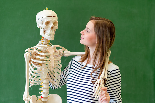 Cool female high school student portrait with an artificial human body skeleton. Student having fun in Biology class. Education concept.