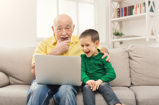 Little boy teaching his grandfather how to use internet safety. Surprised senior man using laptop, laughing with his grandson, sitting on sofa at home, copy space