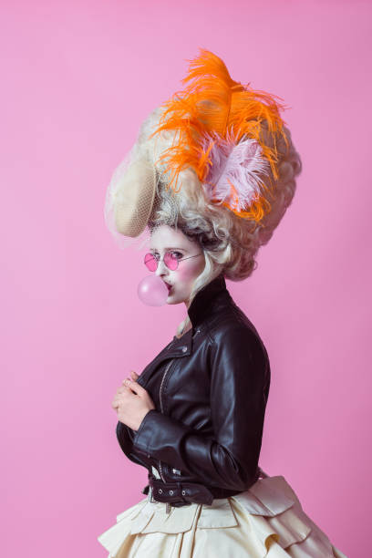 Portrait of rebel woman wearing baroque wig and leather jacket, pink backgroud Portrait of rebel woman wearing a big baroque wig and dress and leather jacket, blowing a bubble gum balloon. Standing against pink background. renaissance dress stock pictures, royalty-free photos & images