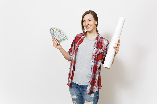 Young smiling beautiful woman in casual clothes holding bundle of dollars, cash money and wallpaper roll isolated on white background. Instruments for renovation apartment room. Repair home concept