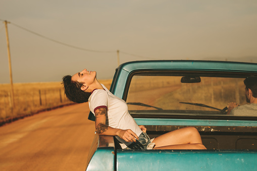 Young woman sitting in the back of a pickup truck enjoying the road trip in country side. Woman relaxing with closed eyes sitting in the car while the man drives the car.
