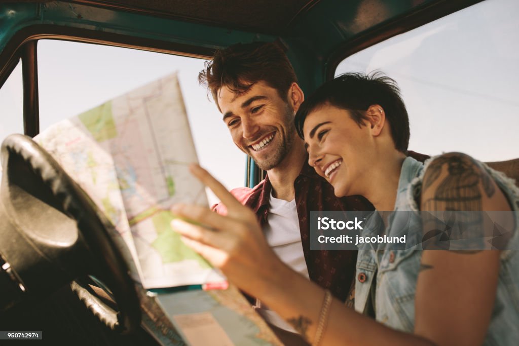 Young travellers on a road trip looking at map Close up of cheerful couple looking at a map sitting in car. Smiling man and woman using map to navigate on a road trip. Road Trip Stock Photo