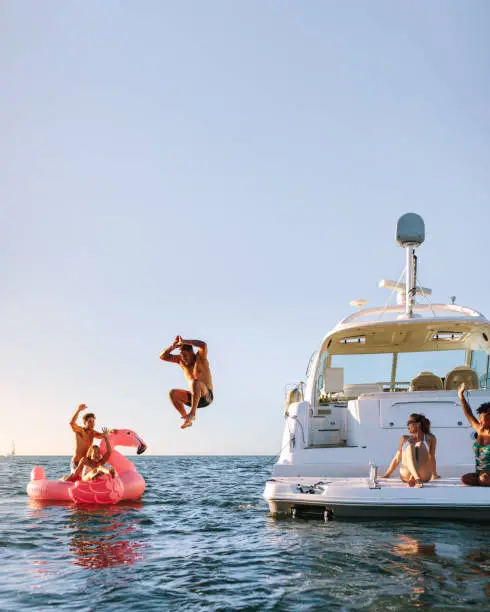 Young man jumping off the boat in to the sea. Young people having fun during party on a private boat. Men and women on yacht and inflatable toy in sea.