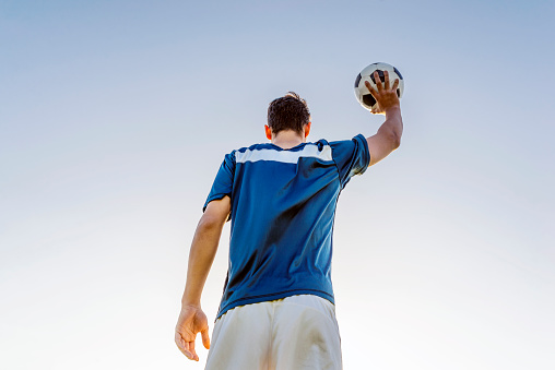 Male soccer player holding a football raising his arm and celebrating after scoring a goal in the stadium against clear blue sky. Rear view  of soccer player is raising arm for the victory during bright sunny day. Photo of Striker football player in blue team celebrating goal in the stadium during match