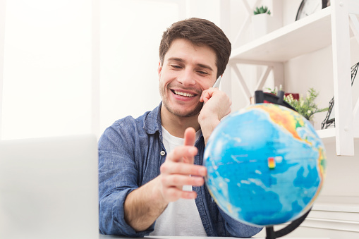 Young happy man playing with globe and talking on phone at home. Handsome guy preparing for vacation, consulting with agent, planning route of trip, booking hotel, copy space